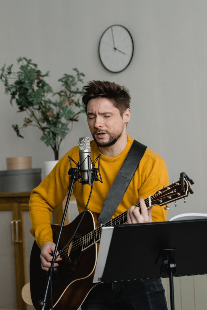 A male musician playing acoustic guitar and singing into a microphone in a cozy indoor setting.